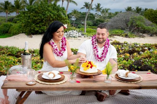 Newlyweds enjoying gourmet tropical meal at outdoor table with Hawaiian leis and lush vegetation