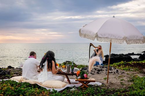 Couple enjoying beach picnic under white umbrella on rocky Hawaii coastline at golden hour