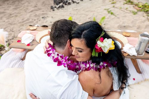 Bride and groom in intimate embrace on sandy beach with pink Hawaiian leis