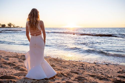 Bride in white wedding dress looking at ocean waves on Hawaii beach at sunset