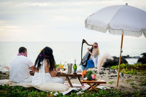 Romantic Hawaii beach picnic setup with white umbrella, cushions, and couple relaxing on rocky shoreline
